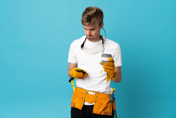 Joven electricista sobre pared aislada sosteniendo café para llevar y un móvil - Foto, Imagen