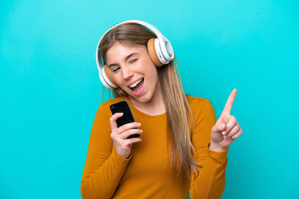 Junge kaukasische Frau isoliert auf blauem Hintergrund Musik hören mit einem Handy und singen - Foto, Bild