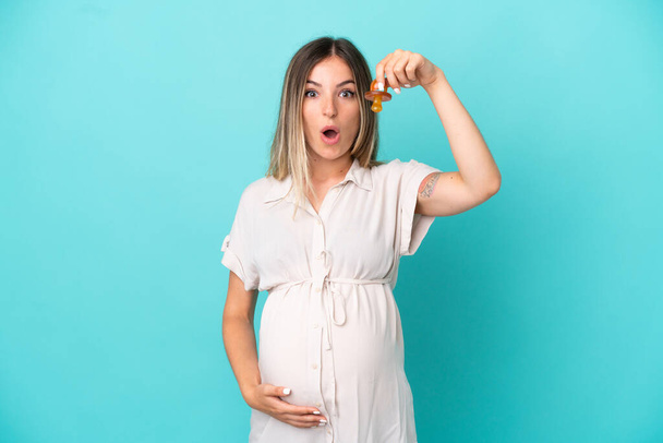 Młoda Rumunka odizolowana na niebieskim tle w ciąży i trzymająca smoczek ze zdumionym wyrazem twarzy - Zdjęcie, obraz