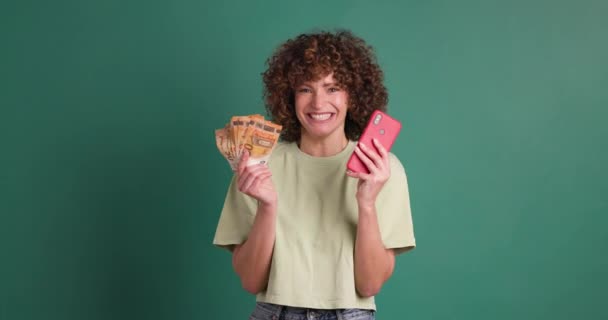 Uśmiechnięta młoda kobieta z kręconymi włosami trzymająca pieniądze i korzystająca z wygranej na smartfonie loterii na zielonym tle - Materiał filmowy, wideo
