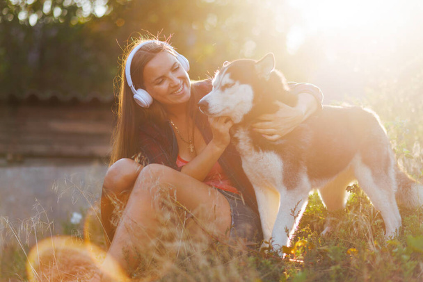 Uma jovem alegre com fones de ouvido abraça seu Husky siberiano em um ambiente natural iluminado pelo sol, compartilhando um momento de relaxamento. - Foto, Imagem