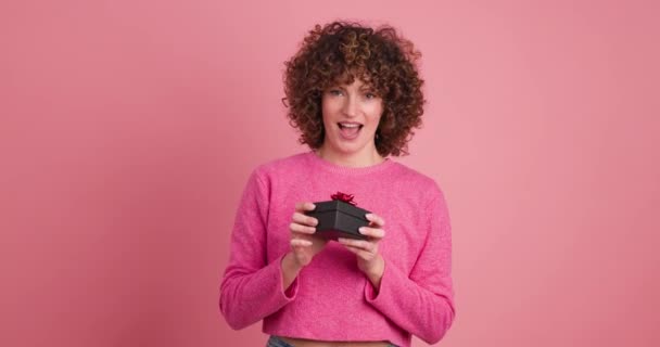 Cheerful young female in pink casual clothes receiving present then opening box with astonished face expression on pastel pink background - Footage, Video