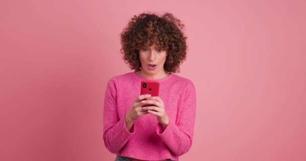 Aufgeregte junge Frau in pinkfarbener Freizeitkleidung mit lockigem Haar checkt Smartphone und feiert gute Nachrichten vor pastellfarbenem Hintergrund - Filmmaterial, Video