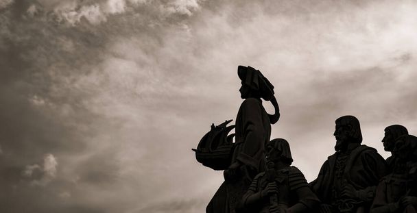 Silhouettes rétroéclairées de sculptures en calcaire et Henry le Navigateur tenant dans sa main un navire du Monument aux Découvertes à Lisbonne avec un ciel nuageux spectaculaire au coucher du soleil. - Photo, image