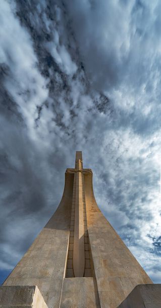 Die flache Frontansicht des Denkmals der Entdeckungen in Lissabon mit einem spektakulären wolkenverhangenen blauen Himmel und dem Abendlicht, das das Steinschwert der Avis-Dynastie beleuchtet. - Foto, Bild