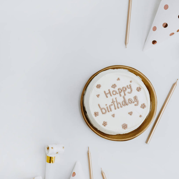 "İyi ki doğdun" yazılı zarif doğum günü pastası, mumlar, beyaz arka planda şenlik konileri. Estetik bayram kutlaması konsepti. Düz yerde, üst tarafta. Beyaz ve altın renkleri - Fotoğraf, Görsel