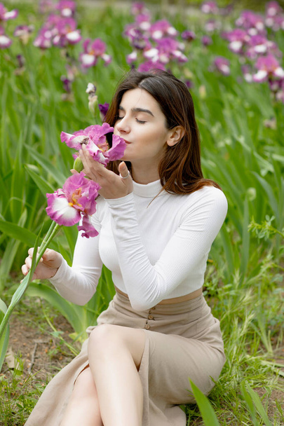 エレガントで若い女性が地面に座り,花を持ち,目を閉じた匂いを嗅いでいる. 春のコンセプト. - 写真・画像
