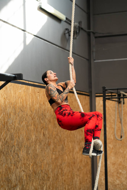 Vertical photo of a strong mature woman climbing a rope in a cross training gym - Photo, Image