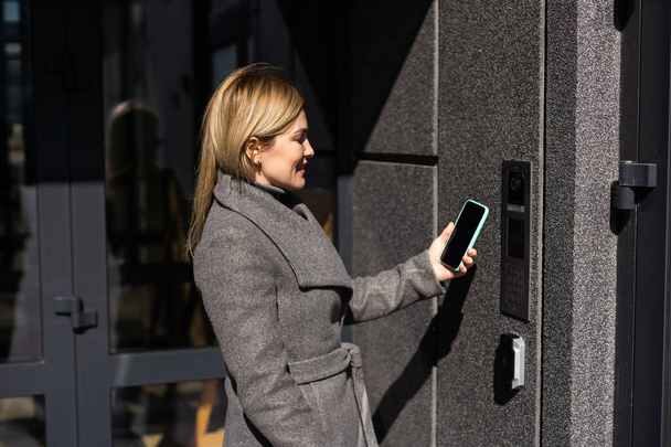 Kobieta blokująca smartlock na drzwiach wejściowych za pomocą smartfona. Koncepcja stosowania inteligentnych zamków elektronicznych z dostępem bez klucza. - Zdjęcie, obraz