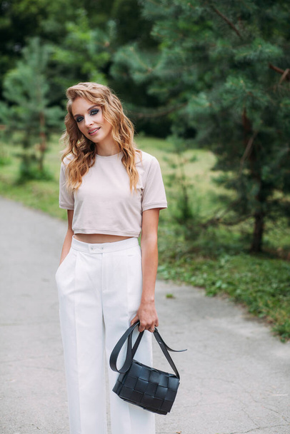 A young slender girl with fashionable make-up and a stylish hairstyle, in white trousers and a beige top, with a black leather handbag in her hands, stands against the background of the park. - Photo, Image