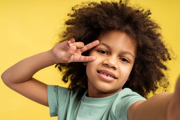 Emociones alegres capturadas. Imagen de una niña afroamericana con el pelo rizado, tomando selfie. Ella mira a la cámara, mostrando el signo de la victoria con los dedos, aislada sobre fondo amarillo - Foto, Imagen