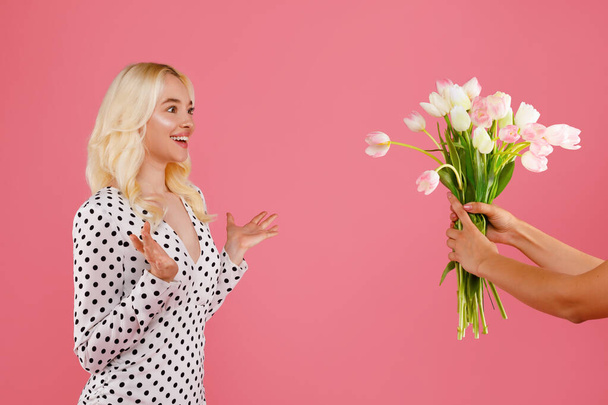 Задоволена блондинка в польковій сукні висловлює несподіванку і радість, оскільки вона збирається отримати прекрасний букет тюльпанів на рожевому фоні для жіночого дня - Фото, зображення