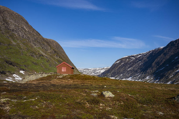De sneeuw bedekte het Breiddalen-dal in het Jotunheimen National Park in Noorwegen, langs de route naar Geiranger, tijdens een zomerdag. Een cabine rust aan de voet van een berg.  - Foto, afbeelding