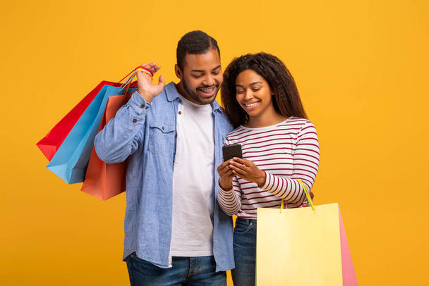 Alışveriş Uygulaması. Afro-Amerikan çift alışveriş torbaları ve akıllı telefon kullanarak mutlu siyah eşler indirimli ve indirimli cep telefonu başvurularını tarıyor ve sarı arka planda duruyor. - Fotoğraf, Görsel