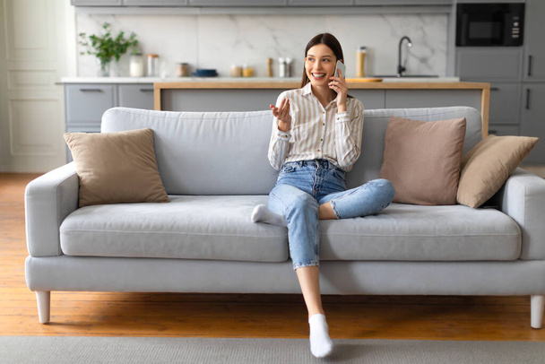 Mujer joven y relajada con camisa de rayas casuales y vaqueros disfrutando de la conversación en el teléfono inteligente mientras se sienta cómodamente en el sofá gris en la sala de estar, espacio libre - Foto, imagen