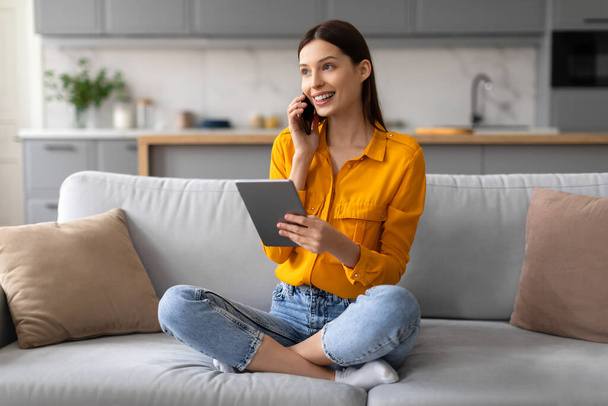 Cheerful young woman comfortably sitting on sofa, simultaneously engaging with digital tablet and talking on smartphone, managing her tasks with ease at home - Photo, Image