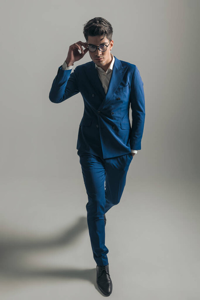 zelfverzekerde jongeman in drievoudig blauw pak die de bril aanpast, hand in hand houdt en voor een grijze achtergrond stapt - Foto, afbeelding