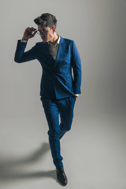 sexy élégant homme en costume bleu regardant sur le côté et de fixer des lunettes tout en marchant avec la main dans la poche en face de fond gris - Photo, image