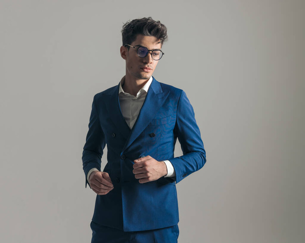 portrait d'un bel homme confiant en costume bleu avec des lunettes regardant loin et posant devant un fond gris - Photo, image