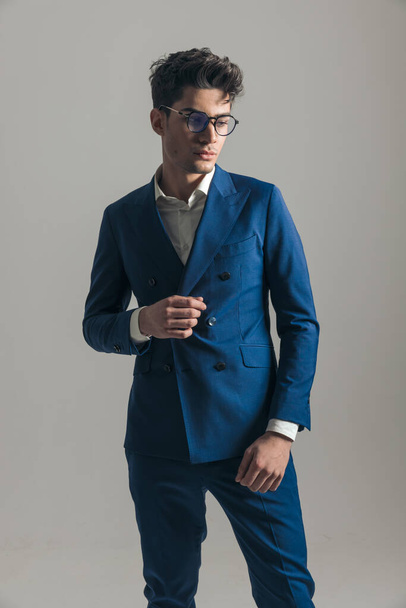 cool stylish businessman with glasses adjusting triple breasted blue suit and looking away in front of grey background - Photo, Image