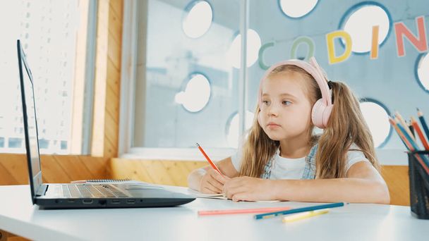 Söpö fiksu tyttö tekee luokkatyötä tai verkko-oppimista kuunnellessaan opettajaa. Onnellinen opiskelija kirjallisesti, piirustus, työskentelevät paperilla, kun yllään kuulokkeet kannettavan tietokoneen ja värikkäitä värikyniä. Virheellisyys. - Valokuva, kuva