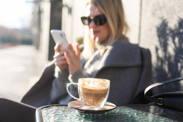 Nuori nainen nauttii kahvia, istuu matkapuhelimen kanssa kahvilan terassilla vanhan kaupungin kadulla aurinkoisena päivänä. Laadukas kuva - Valokuva, kuva