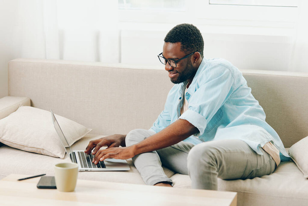 笑顔のアフリカ系アメリカ人男性は,現代のリビングルームでソファーに座っている間,ノートパソコンで作業し,電話でオンラインショッピングとコミュニケーションに従事 このアップビートフリーランサーはプロフェッショナリズムを誇っています - 写真・画像