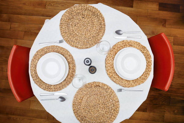 Draufsicht auf einen runden Esstisch mit weißen Tellern, Besteck, gewebten Tischsets und roten Stühlen, bereit für eine Mahlzeit. - Foto, Bild