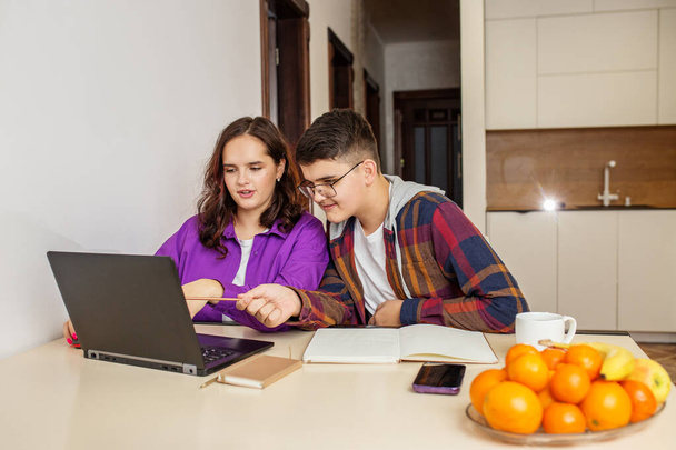 Dwóch nastolatków, dziewczyna i chłopiec, angażują się w sesję grupową z laptopem, książkami i papeterią na stole kuchennym. - Zdjęcie, obraz