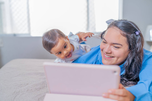 Un bebé alegre se acerca mientras disfruta de una tableta digital con su madre, compartiendo un momento de vinculación. - Foto, imagen