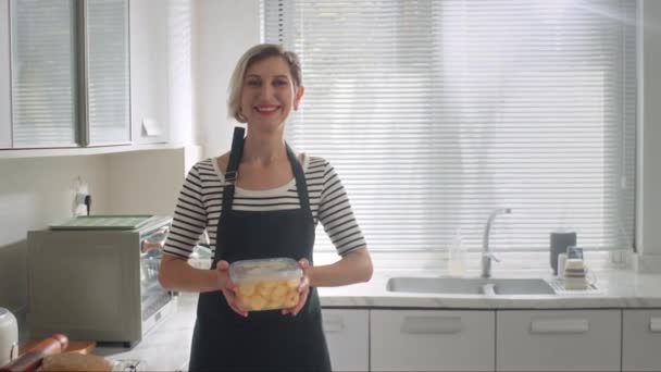 Ritratto di donna caucasica positiva che tiene un contenitore con pasta per biscotti in cucina - Filmati, video