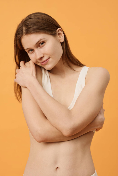 Vertical medium studio portrait of young Caucasian woman with no makeup posing for camera hugging herself and looking at camera - Photo, Image