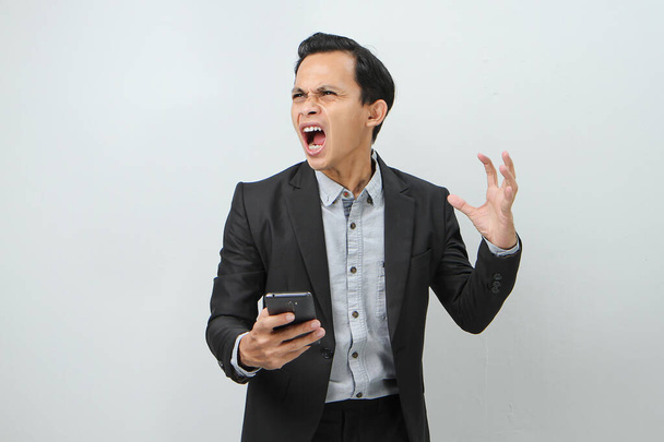 angry mad asian indonesian business man in suit holding smart phone on isolated background - Photo, Image