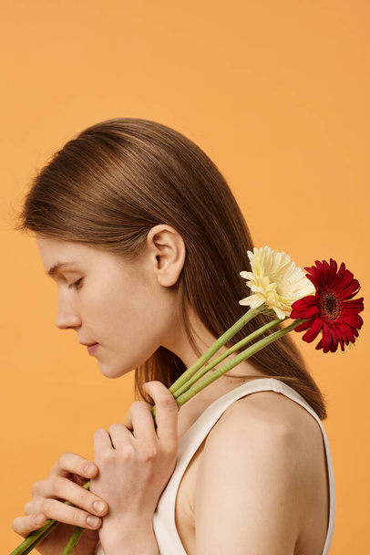 新鮮なゲルベラの花を持つカメラのためのメイクなしの若い白人女性の縦の側面の眺めの中型のクローズアップスタジオショット - 写真・画像