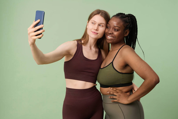 Mittleres Studioporträt zweier junger, ethnisch unterschiedlicher Frauen in Sportbekleidung, die ein Selfie mit der Smartphone-Kamera machen - Foto, Bild