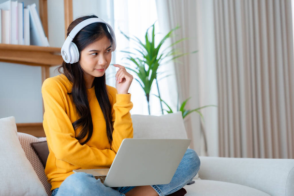 Fiatal, imádnivaló ázsiai nő mosolyog, fülhallgatót visel, otthon dolgozik a nappaliban. Boldog gyönyörű nő számítógéppel laptop és ül a kanapén otthon, Videó hívás barátjával - Fotó, kép