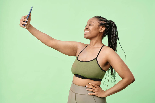 Ritratto medio della giovane donna nera moderna con i capelli intrecciati che indossa activewear in piedi in studio in posa per foto selfie - Foto, immagini
