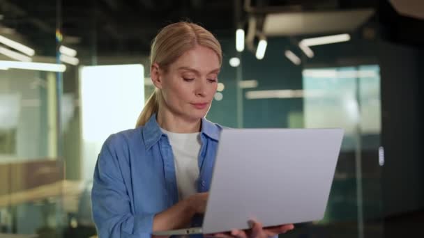Konsantre olmuş yetişkin bir kadın gri dizüstü bilgisayarı elinde tutuyor ve camdan çalışma alanına bakıyor. Özenli yönetici verimli çalışıyor ve sorunların çözümünü kendine güvenen bir gülümsemeyle buluyor. - Video, Çekim