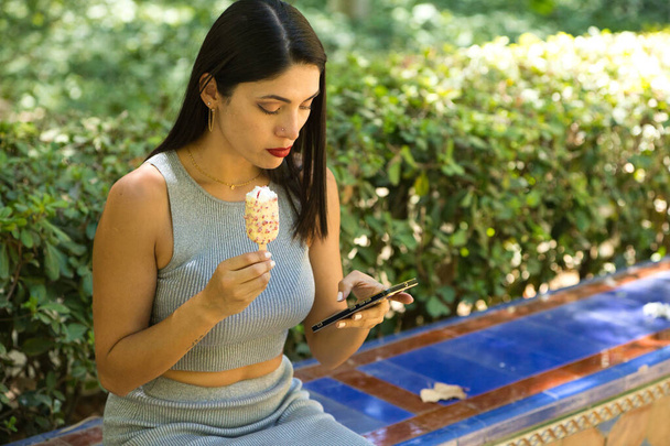 Latina, junge und schöne Brünette, zückt ihr Handy und isst ein erfrischendes Eis, um die hohen Temperaturen in der Stadt zu bekämpfen. Hitzewelle und Sommerkonzept - Foto, Bild