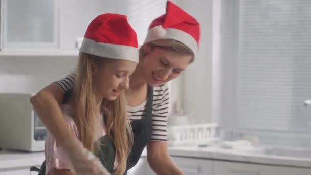 キッチンで一緒に料理するクリスマスクッキーのために彼女の娘にひざまずいた生地を教える母親のショットを傾けて - 映像、動画