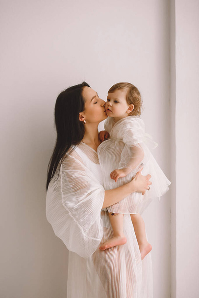 Studio portret van een gelukkige moeder in witte jurk die naast de muur staat en haar dochtertje in haar armen houdt en kust. Moeder en dochter in witte kleren. Het concept van gelukkig moederschap - Foto, afbeelding