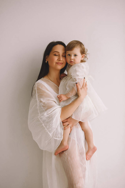 Studio portret van een gelukkige moeder in witte outfit die naast de muur staat en haar dochtertje in haar armen houdt. Moeder en dochter in witte jurken. Het concept van een gelukkige jeugd. Kopieer ruimte voor tekst - Foto, afbeelding