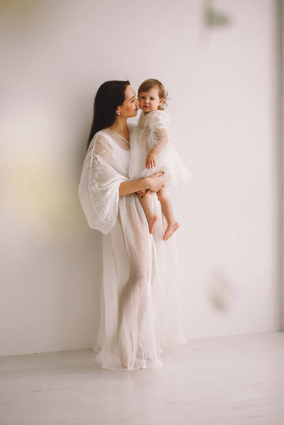 Onnellinen äiti valkoisessa asussa seisoo lähellä seinää studiossa ja pitää tytärtään sylissään. Äiti ja tytär valkoisissa mekoissa. Onnellisen lapsuuden käsite. Kopioi tilaa tekstille - Valokuva, kuva