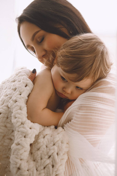 Mère heureuse en tenue blanche embrassant sa petite fille à la maison. Petite fille enveloppée dans une couverture tricotée à la maison. Protection parentale, soins et amour. Mère et fille - Photo, image