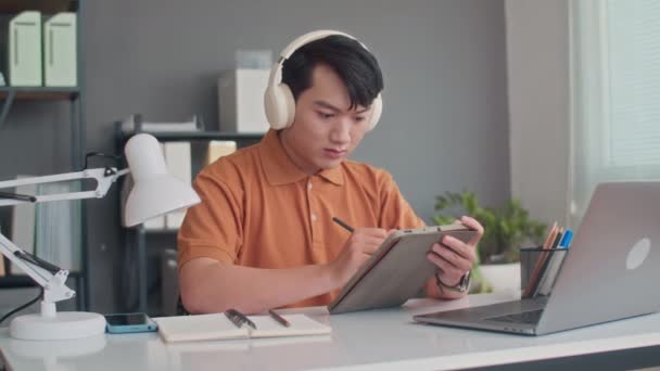 Fotografía media de un joven desarrollador asiático en la aplicación de prueba de auriculares en la tableta digital y el portátil en la oficina - Metraje, vídeo