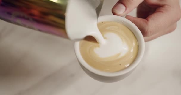 Barista dělá cappuccino a nalévá mléčnou pěnu do nápoje ve tvaru srdce. Koncept latté. Oříznutý pohled na muže připravující čerstvý kofeinový nápoj v kavárně  - Záběry, video