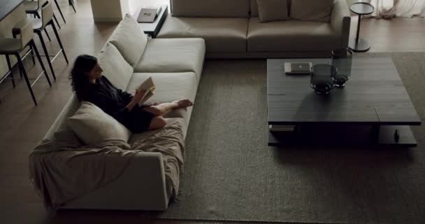 Žena sedí na pohodlném gauči v obývacím pokoji, obklopena dřevěným nábytkem a podlahou, čte si knihu - Záběry, video