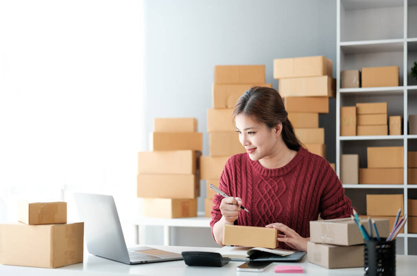 Uruchomienie małych przedsiębiorstw MŚP przedsiębiorca niezależny azjatycki kobieta noszenie fartuch za pomocą laptopa i pudełka do odbierania i przeglądu zamówień online, aby przygotować się do pakowania sprzedaży klientom, online sme pomysłów biznesowych - Zdjęcie, obraz