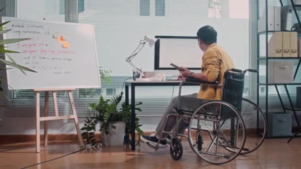Довгий знімок, вид ззаду пустотливого чоловічого кодера в інвалідному візку, що працює на комп'ютері в офісному столі - Кадри, відео