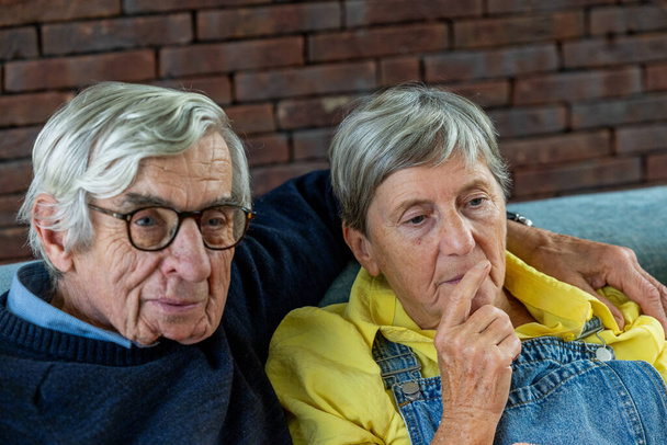 Esta imagem fornece uma captura sincera de um casal de idosos em um momento de reflexão. Sentado confortavelmente contra um pano de fundo de parede de tijolo que adiciona um toque de charme rústico, o cavalheiro está vestido com um - Foto, Imagem